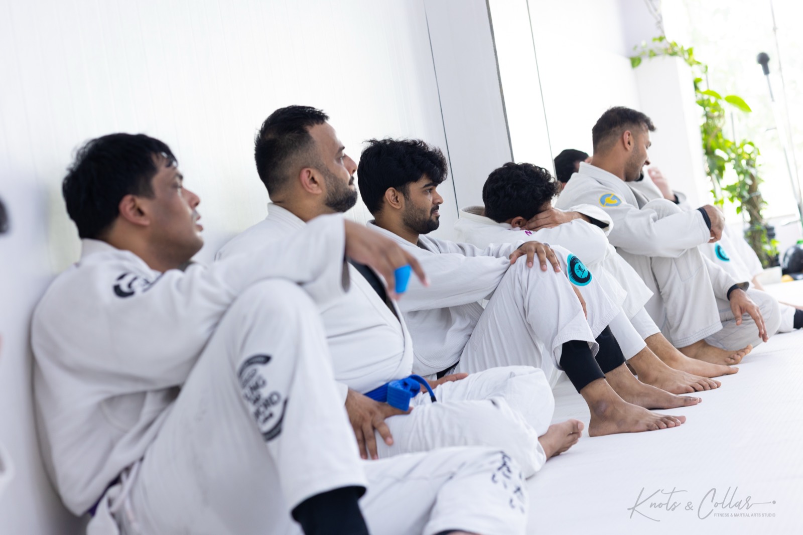 A row of adult jiu-jitsu athletes in white gis with blue and black belts sitting mat-side watching a match