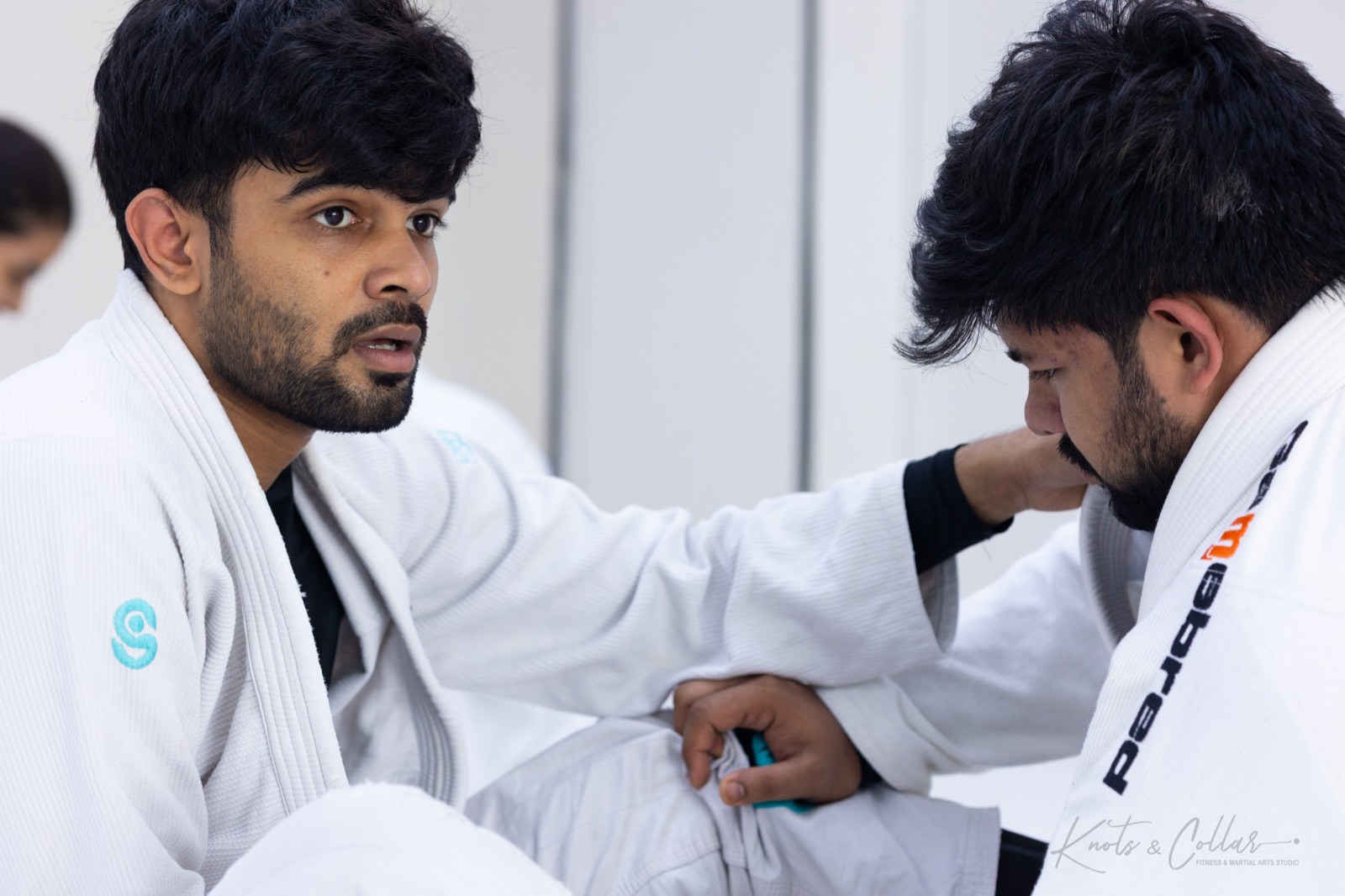 Two adult jiu-jitsu teammates in white gis talking on the mat between rounds at a Delhi training session