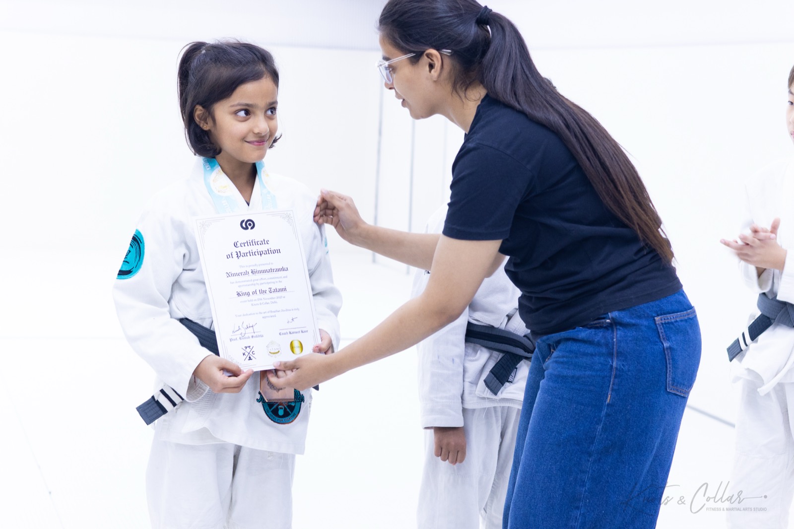 A coach presenting a certificate and medal to a young girl in a white gi at a kids jiu-jitsu tournament